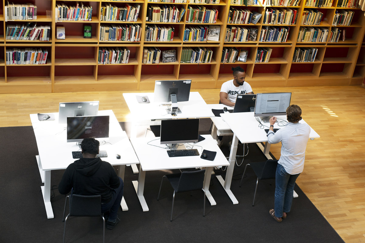 Datorer vid höj- och sänkbara bord. Längs ena väggen finns böcker i bokhyllor. Två personer sitter vid varsin dator. En person står vid en dator.