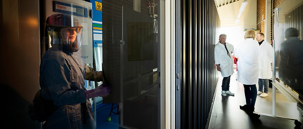 Skyddsklädd person bakom glasdörr i laboratorium. Utanför står tre forskare i vita rockar.