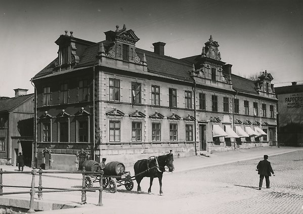 Black and white photo of the Theatrum Oeconomicum on Old Square. Outside, two black-clad people in hats and a horse and carriage walk. Next to it stands a wooden house.