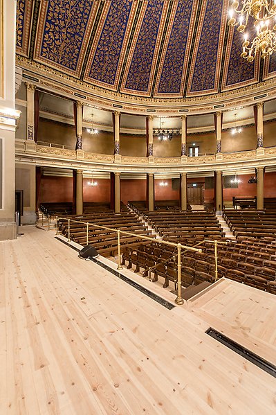 The left side of the grand auditorium as seen from the center of the stage. The draperies for the balcony seats at the back are gone. The balcony seats form a semicircle and are supported by rows of columns. A loudspeaker stands on the stage floor.